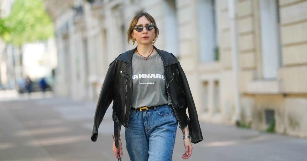 PARIS, FRANCE - APRIL 09: Emy Venturini wears a black leather jacket with wide lapels and zipped pockets, layered over a faded grey graphic T-shirt by Zara featuring “WANNABE” and the Spice Girls logo. Blue high-waisted jeans by The Frankie Shop and a black belt with a gold H buckle by Hermes are visible. Gold sculptural earrings by Rabanne and a red beaded necklace complete the look. Mid shot, during a street style fashion photo session, on April 09, 2025 in Paris, France. (Photo by Edward Berthelot/Getty Images)